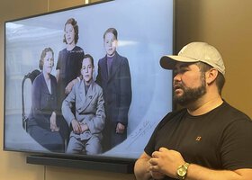 Mann med skjegg og caps snakker til noen foran en presentasjon av et gammelt familiefotografi - Klikk for stort bilde